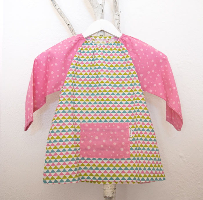 Bata de triángulos en rosa, verde y azul Bata para guardería, con estampado de triángulos y mangas y bolsillo delantero en rosa.